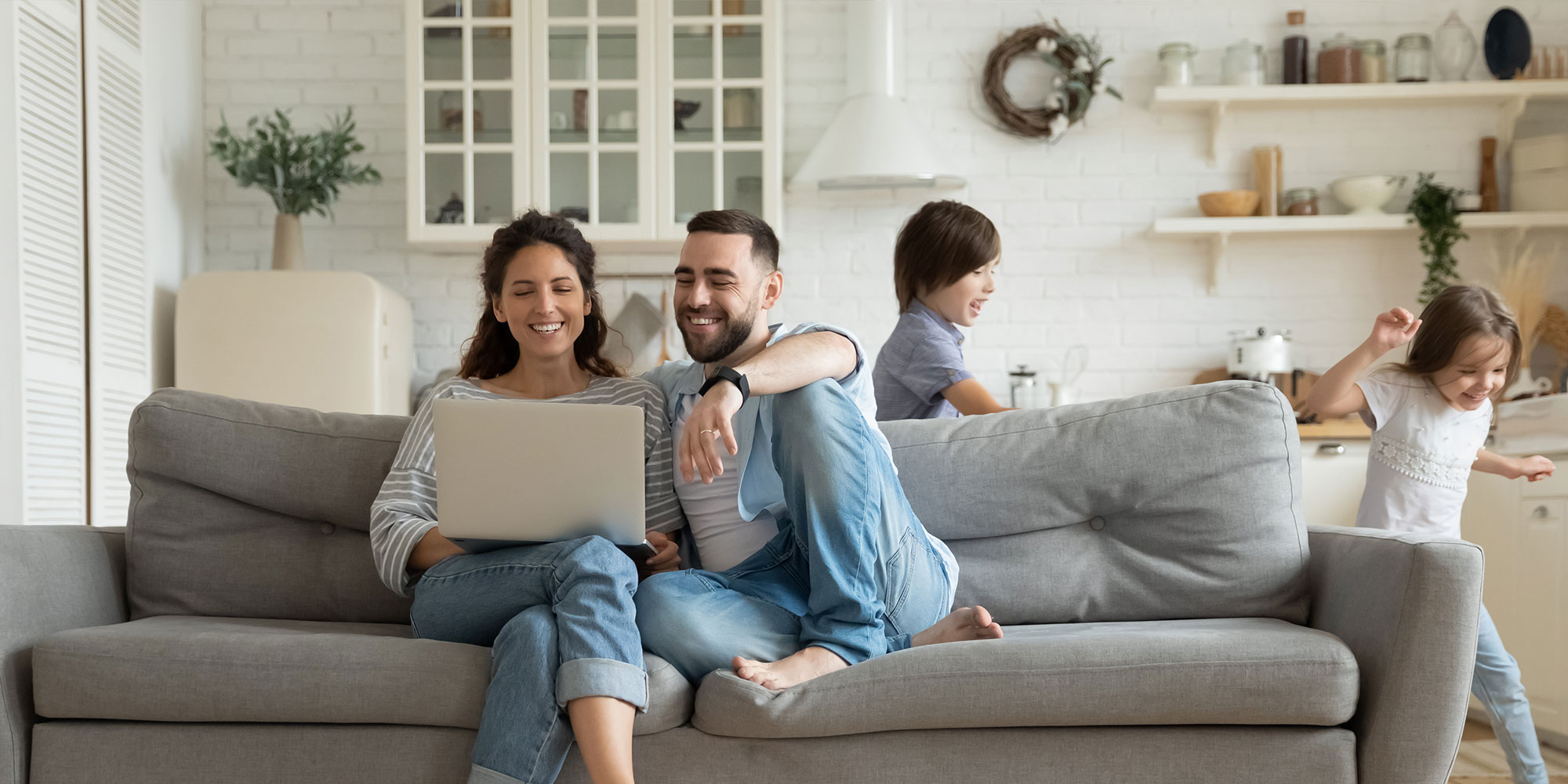 A couple sits on a couch looking at a laptop while two children play behind